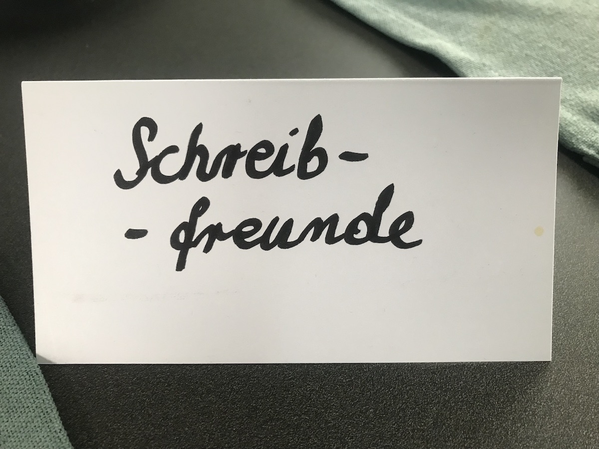 Weißer, zu einem "Aufsteller" gefalteter Karton mit dem handschriftlichen Text "Schreibfreunde"