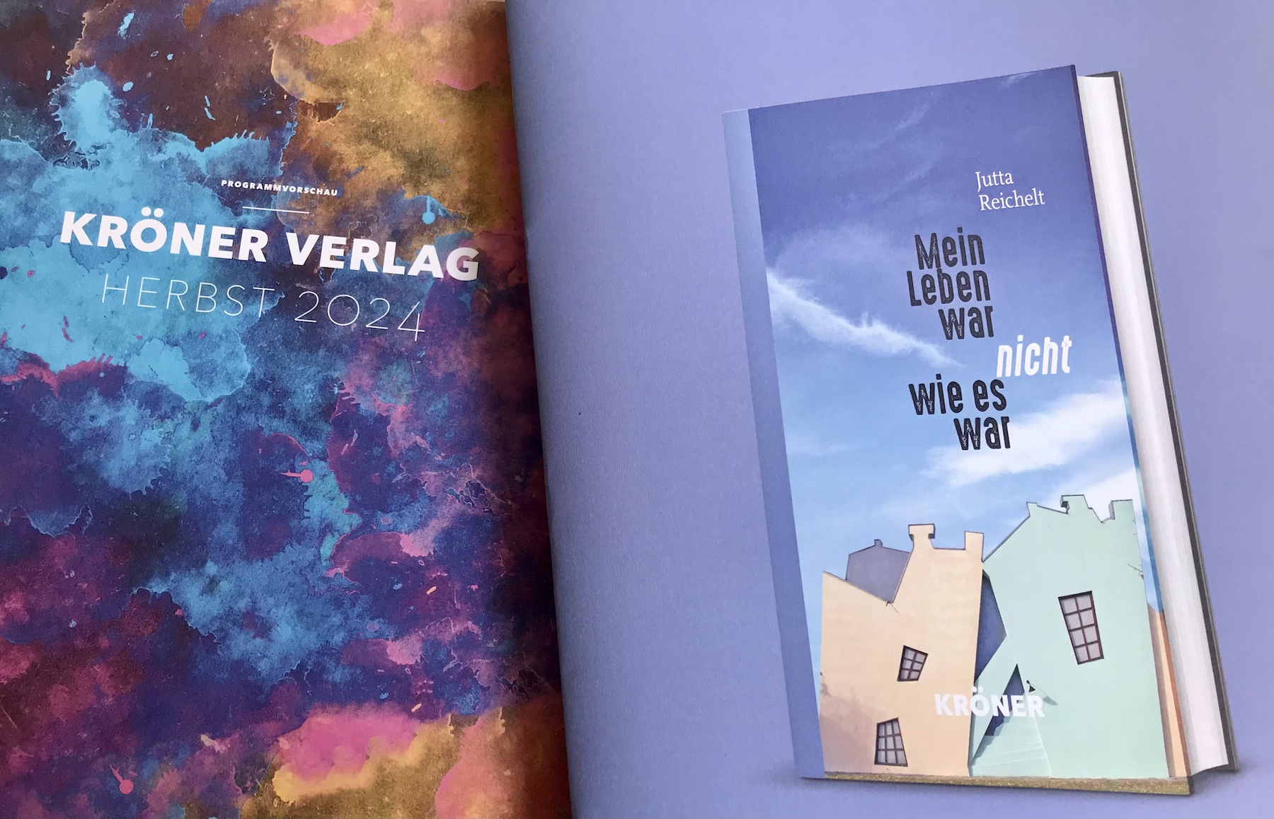 Auf der linken Bildhälfte das Cover der Herbstvorschau 2024 des Kröner Verlags auf der rechten Hälfte ein Buch-Cover mit dem Text: Jutta Reichelt und "Mein Leben war nicht, wie es war!" Der Text befindet sich auf blauem Hintergrund (Immer), darunter sind zwei Fassaden zu sehen, an denen alles schief und seltsam "verrückt" erscheint.